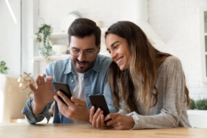 Young happy couple using two phones share social media news at home, smiling husband and wife millennial users customers talking doing shopping online sit at table, mobile tech lifestyle concept